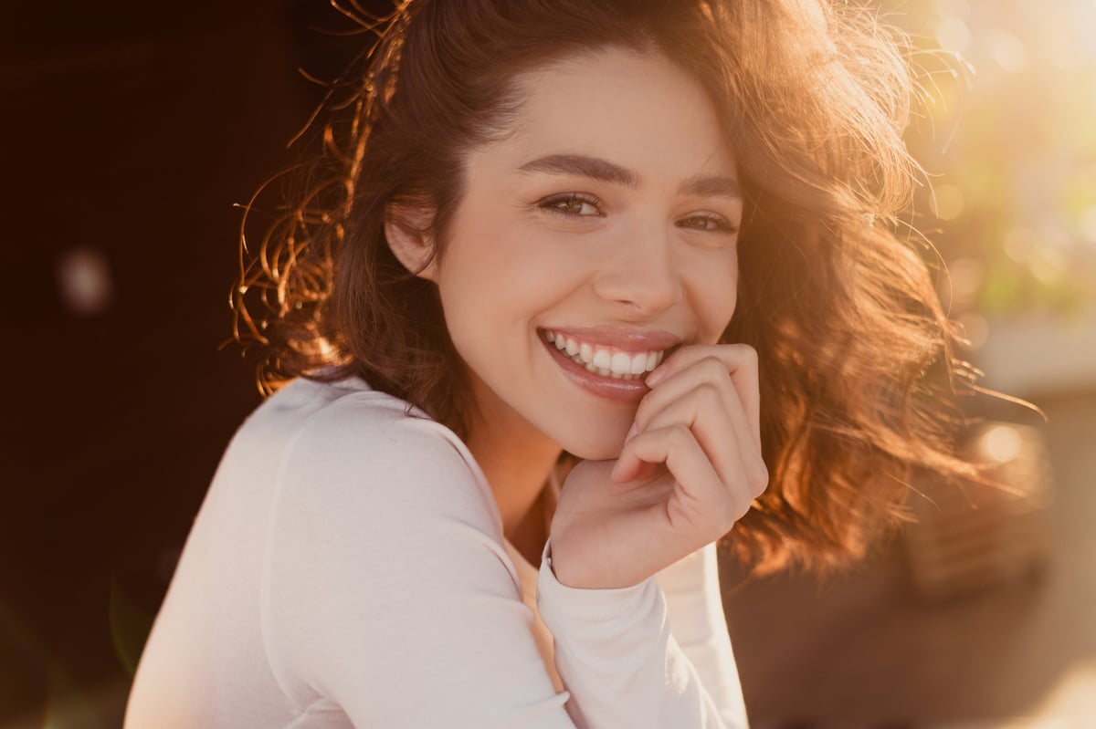 Young woman smiles in a close-up portrait bathed in warm golden-hour sunlight, conveying natural beauty, joy and relaxed confidence suitable for lifestyle, wellness, beauty and personal care concepts.